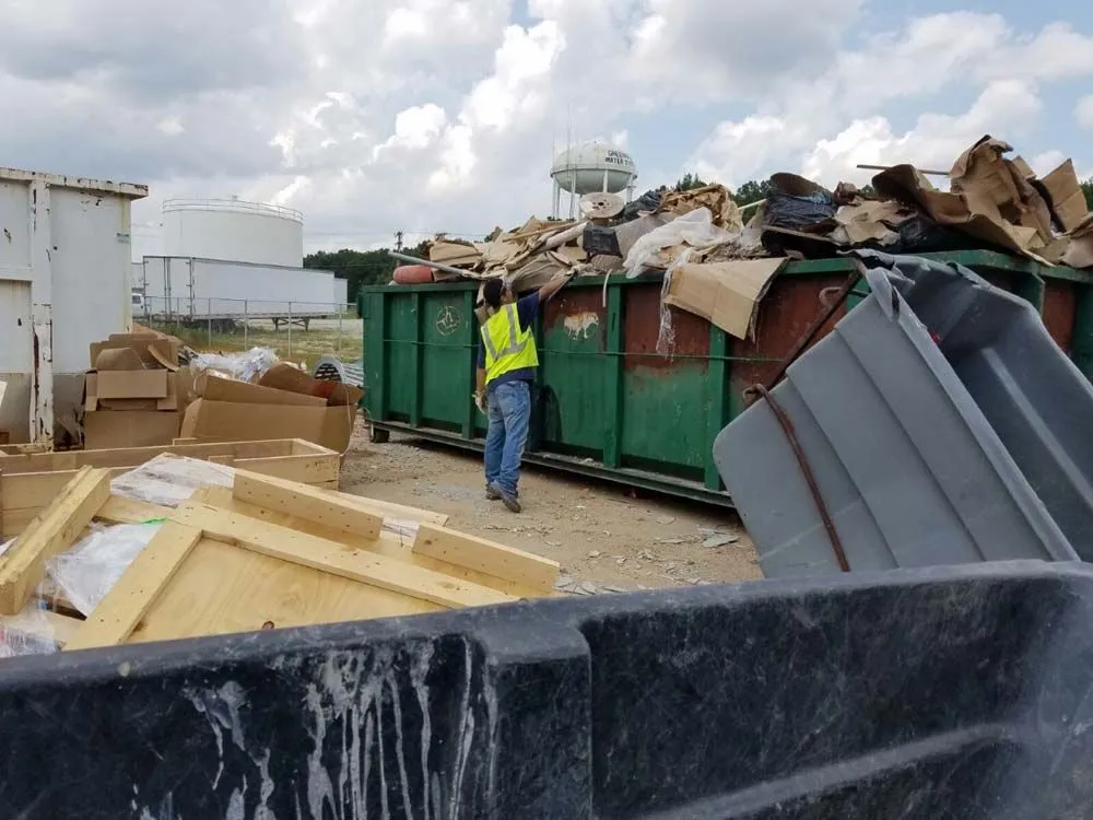 A man in a yellow vest is standing in front of a dumpster filled with cardboard boxes.
